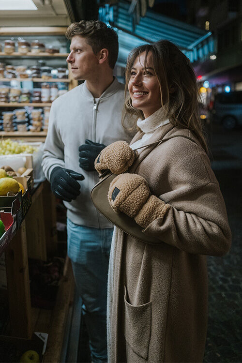 Junge Frau mit beigen Fäustlingen aus Fleece-Material lächelt an einem Marktstand, daneben steht ein Mann mit schwarzen Handschuhen.