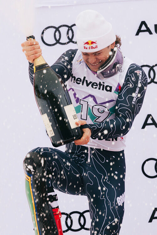 Skirennläufer im Wettkampfanzug sprüht mit einer großen Champagnerflasche auf dem Siegerpodest.“
