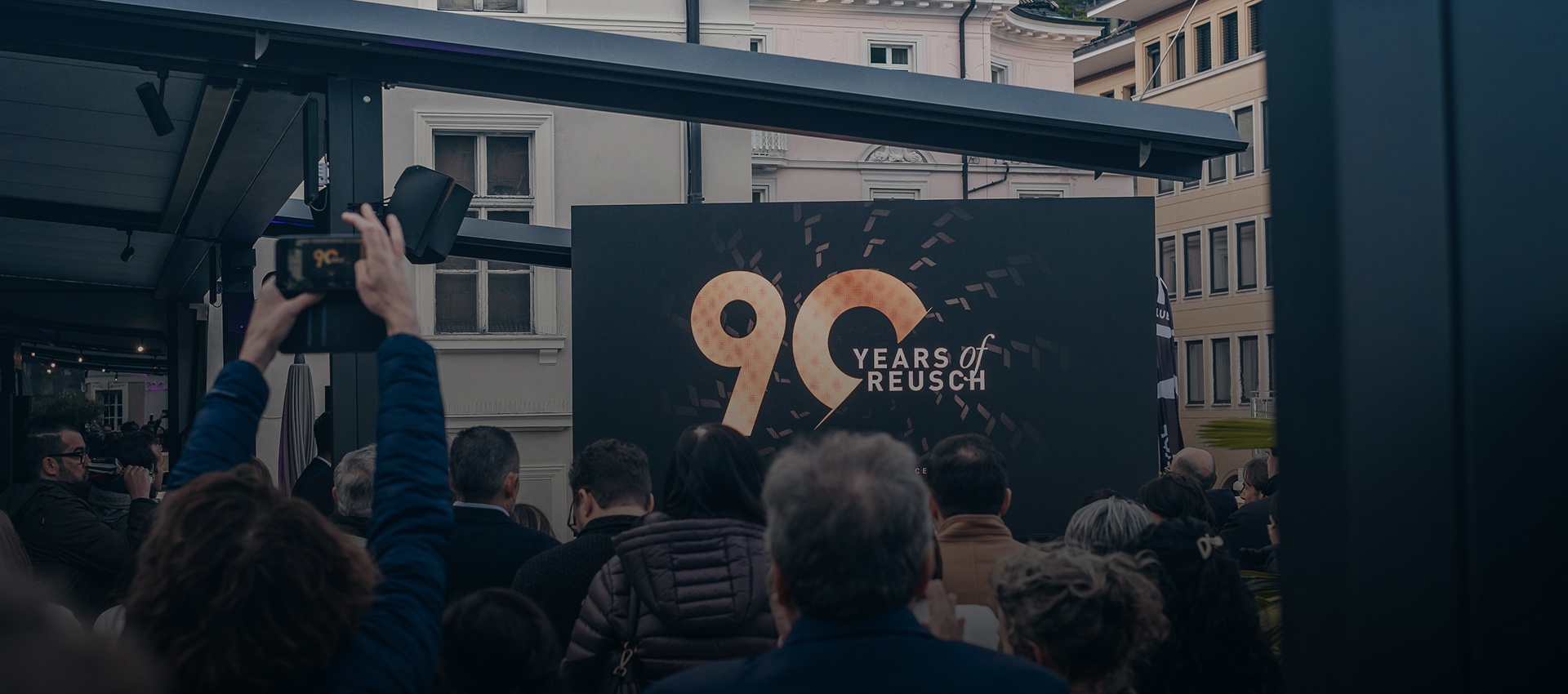 Menschenmenge bei einer Veranstaltung zum 90-jährigen Jubiläum von Reusch, mit großem Bildschirm im Hintergrund, der das Logo ‚90 Years of Reusch‘ zeigt.